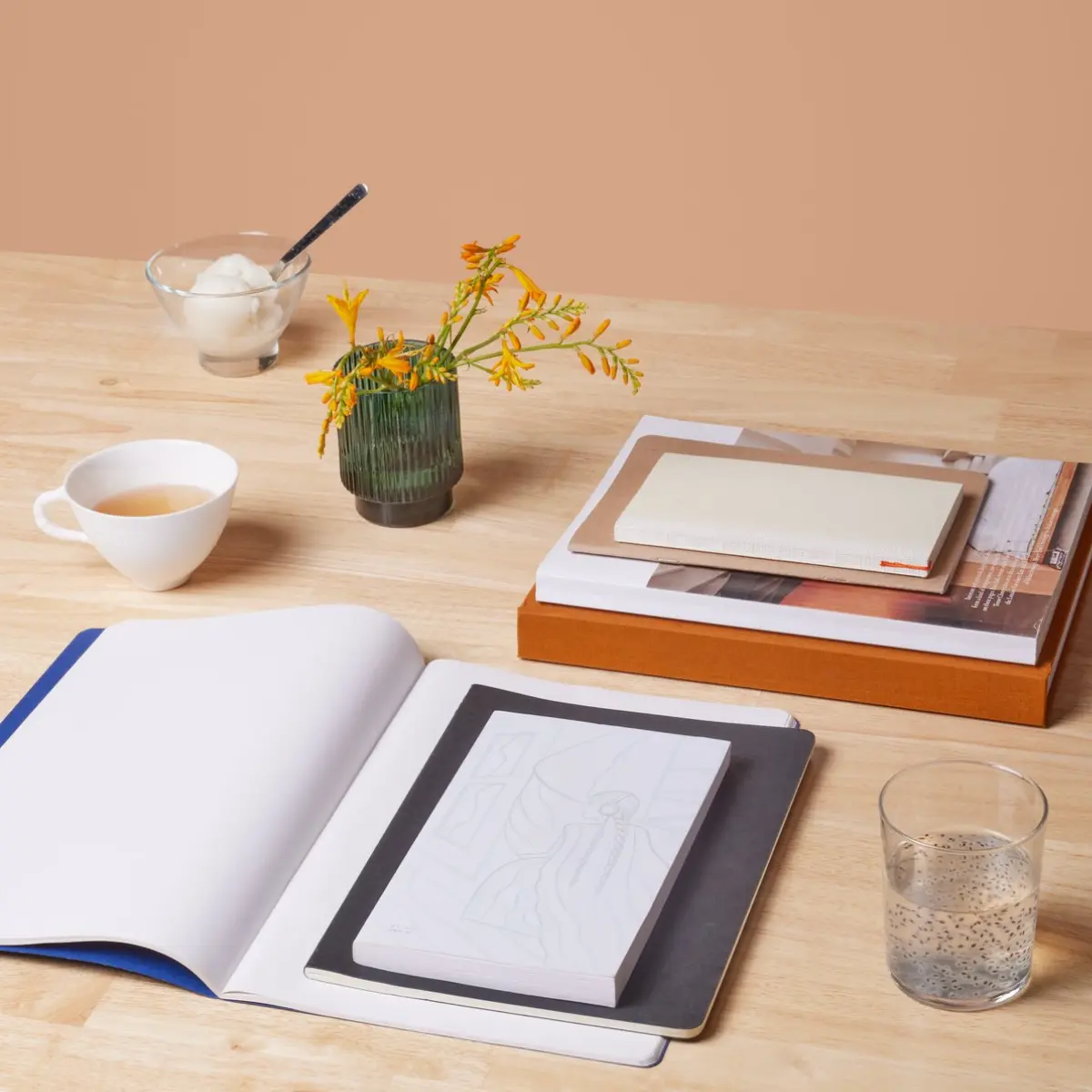 Pencils and a few natural pebbles are scattered nearby on a light wooden table. The lighting is soft and focused on the Boznańska artwork and paper, highlighting the texture and creative process.